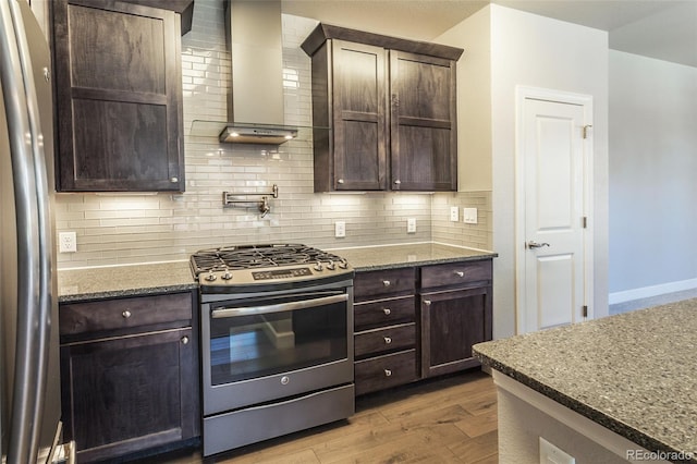 kitchen featuring wall chimney exhaust hood, stainless steel appliances, light hardwood / wood-style flooring, dark stone counters, and decorative backsplash