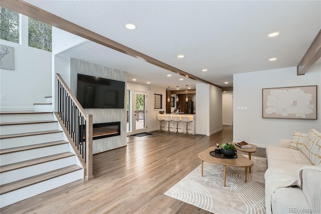 living area featuring beam ceiling, recessed lighting, stairway, a glass covered fireplace, and wood finished floors