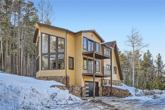 snow covered house featuring a balcony, stone siding, and a garage