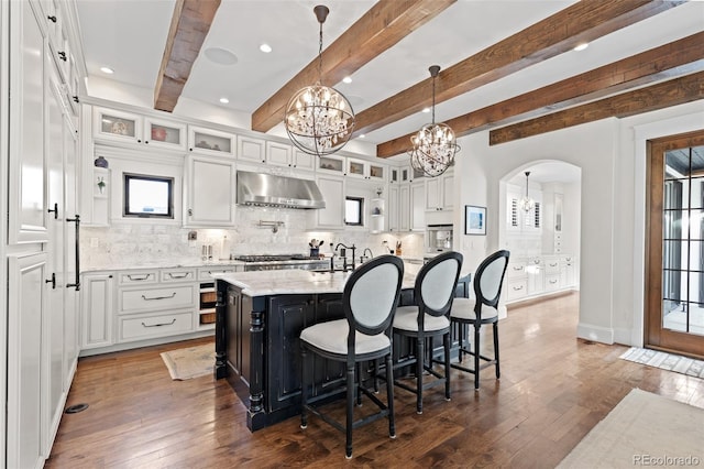 kitchen with a chandelier, dark wood-type flooring, a kitchen island with sink, under cabinet range hood, and white cabinetry