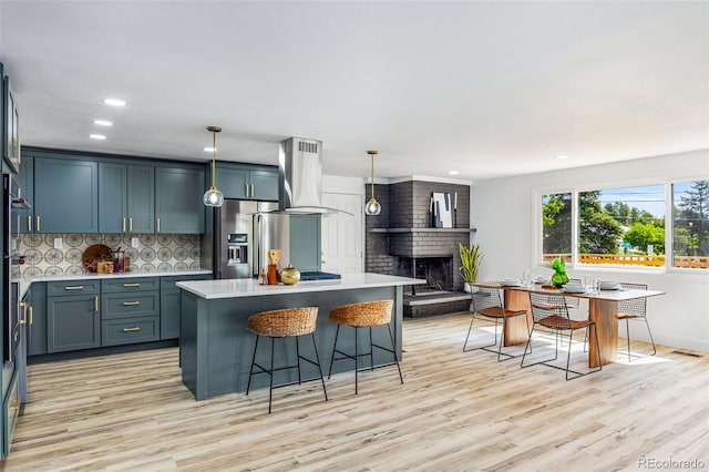 7120 S Sheridan Ct, Littleton, CO 80128 - kitchen featuring island exhaust hood, stainless steel refrigerator with ice dispenser, light wood-style flooring, decorative backsplash, and a brick fireplace