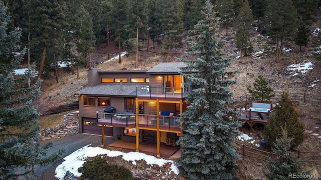 back of house with aphalt driveway, a deck, a balcony, a view of trees, and a garage