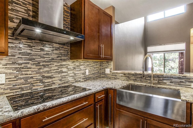 kitchen featuring wall chimney range hood, light stone countertops, a sink, black electric cooktop, and decorative backsplash