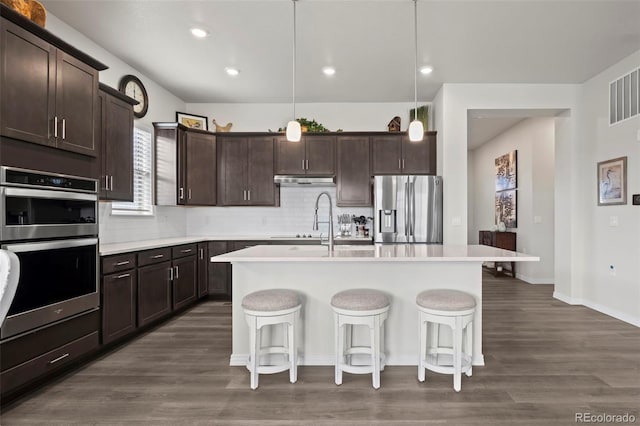 kitchen with dark brown cabinetry, stainless steel appliances, decorative light fixtures, and an island with sink