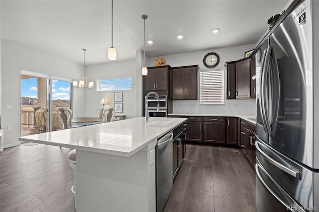 kitchen featuring dark wood-type flooring, appliances with stainless steel finishes, hanging light fixtures, and a center island with sink