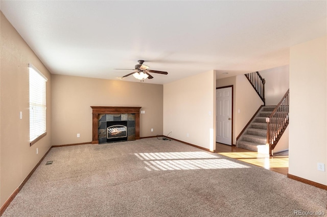 unfurnished living room with baseboards, a tile fireplace, ceiling fan, stairway, and carpet flooring