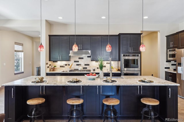 24322 E Mineral Dr, Aurora, CO 80016 - kitchen with under cabinet range hood, stainless steel double oven, tasteful backsplash, light stone counters, and a kitchen bar