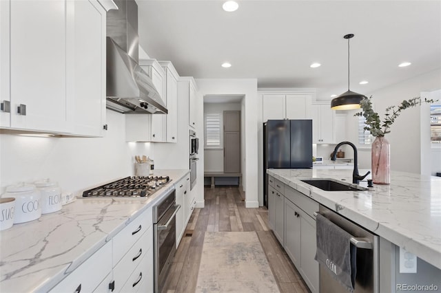 kitchen featuring light wood finished floors, appliances with stainless steel finishes, white cabinetry, a sink, and wall chimney exhaust hood