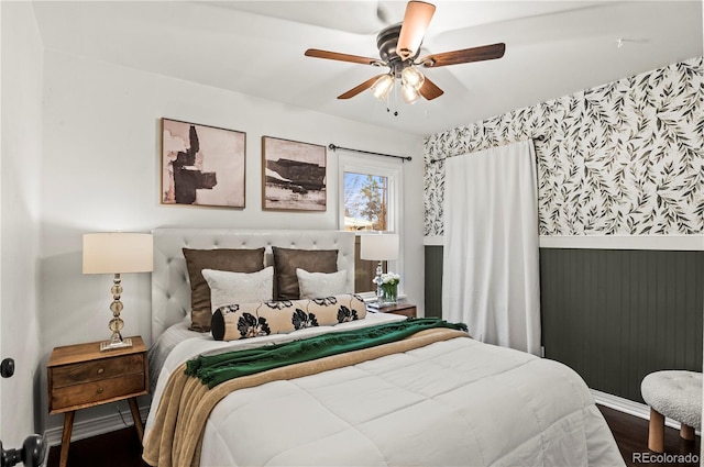 bedroom with wallpapered walls, wood finished floors, a wainscoted wall, and ceiling fan