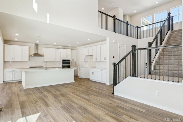 kitchen with white cabinetry, an island with sink, a high ceiling, wall chimney range hood, and light hardwood / wood-style flooring