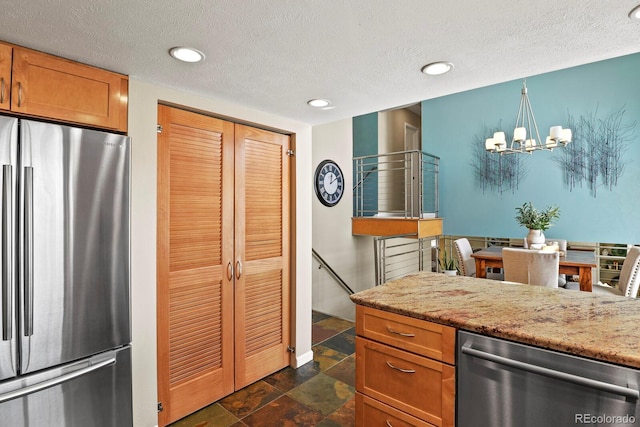 kitchen with brown cabinets, a notable chandelier, stone finish flooring, appliances with stainless steel finishes, and light stone countertops