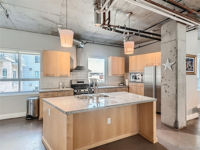 kitchen featuring light brown cabinets, wall chimney range hood, hanging light fixtures, stainless steel appliances, and an island with sink