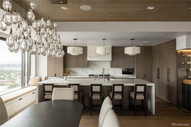 interior space with decorative light fixtures, wood-type flooring, tasteful backsplash, an island with sink, and dark brown cabinetry