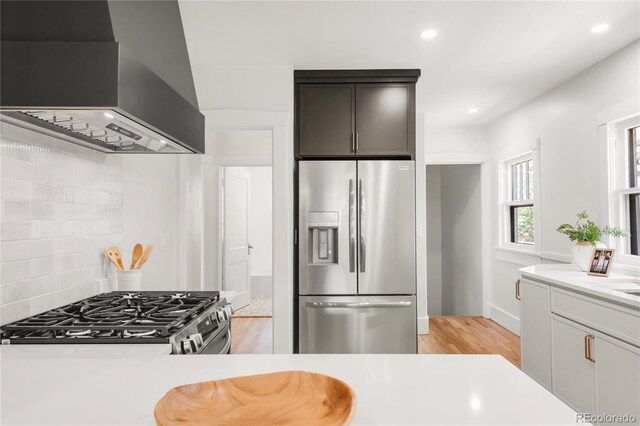 kitchen featuring wall chimney range hood, appliances with stainless steel finishes, tasteful backsplash, light countertops, and light wood-style floors
