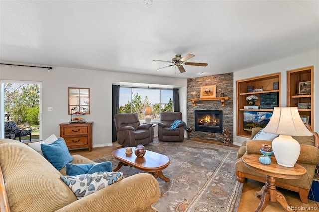 living room featuring a ceiling fan, a stone fireplace, wood finished floors, baseboards, and built in features