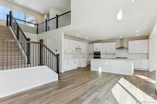 13900 Hanging Lake St, Parker, CO 80138 - kitchen with wall chimney range hood, white cabinets, light wood-style floors, a towering ceiling, and a kitchen island with sink