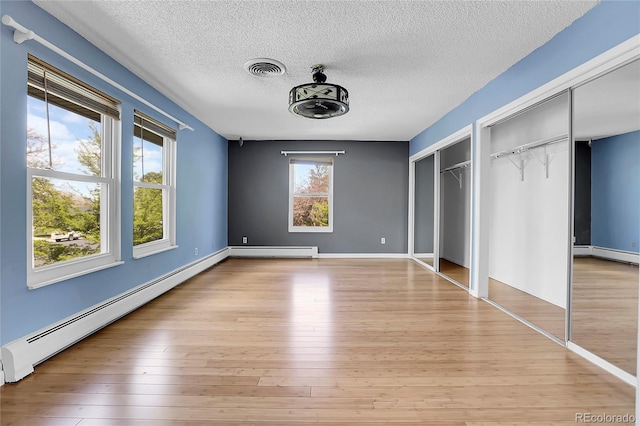 unfurnished bedroom featuring baseboard heating, two closets, a textured ceiling, light wood-type flooring, and baseboards