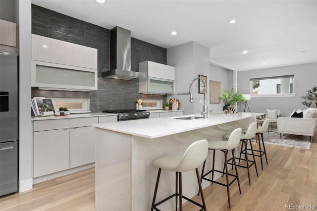 kitchen featuring light countertops, appliances with stainless steel finishes, a kitchen island with sink, a sink, and wall chimney range hood