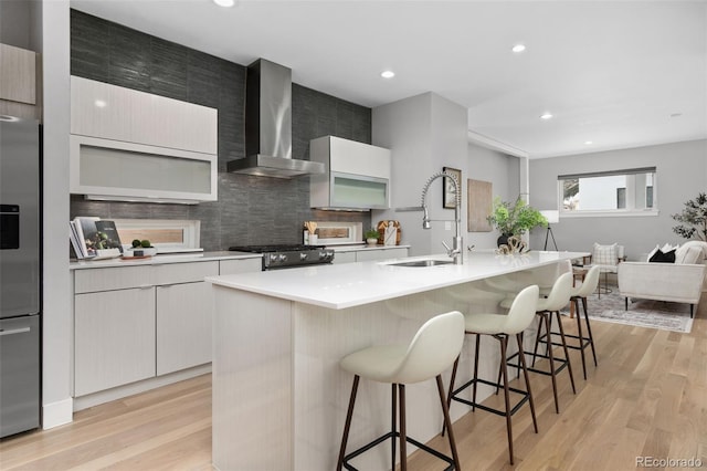 kitchen featuring a sink, wall chimney range hood, appliances with stainless steel finishes, a kitchen bar, and light wood finished floors