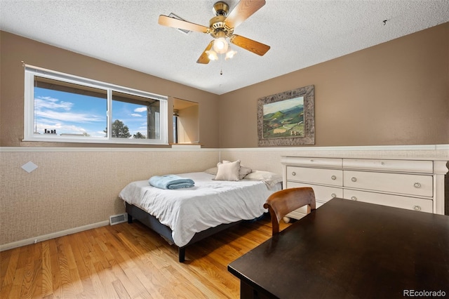bedroom featuring a textured ceiling, a ceiling fan, wainscoting, baseboards, and light wood-style floors