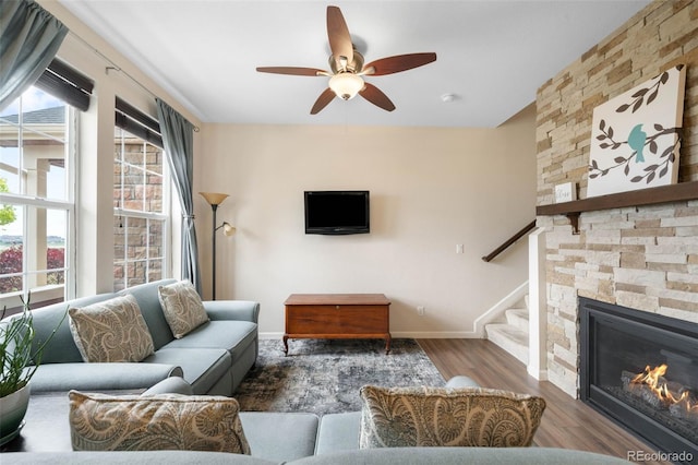 living area featuring stairs, wood finished floors, baseboards, a stone fireplace, and ceiling fan