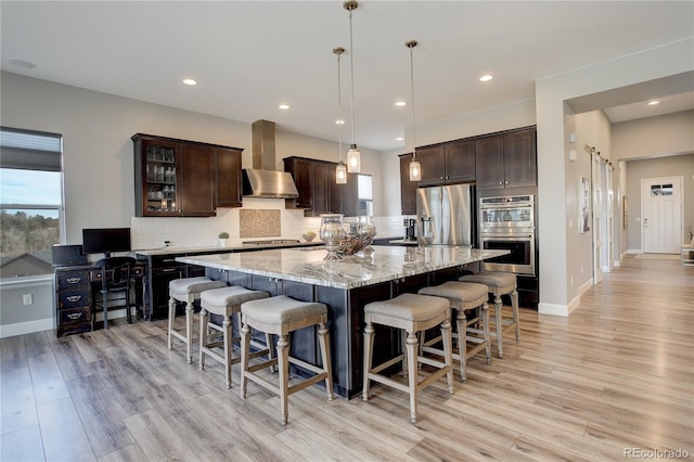 kitchen with appliances with stainless steel finishes, dark brown cabinetry, wall chimney range hood, decorative light fixtures, and a large island