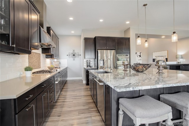 kitchen featuring backsplash, a large island with sink, wall chimney range hood, hanging light fixtures, and stainless steel appliances