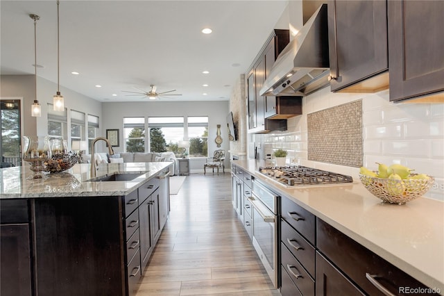 kitchen featuring a center island with sink, sink, wall chimney exhaust hood, decorative light fixtures, and stainless steel appliances