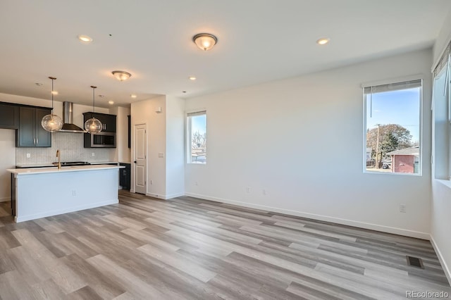 kitchen featuring wall chimney exhaust hood, a kitchen island with sink, decorative light fixtures, decorative backsplash, and light wood-type flooring