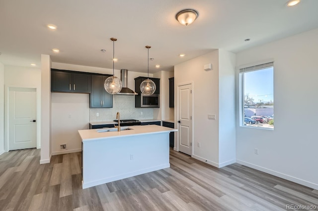 kitchen featuring sink, wall chimney exhaust hood, pendant lighting, a kitchen island with sink, and light wood-type flooring