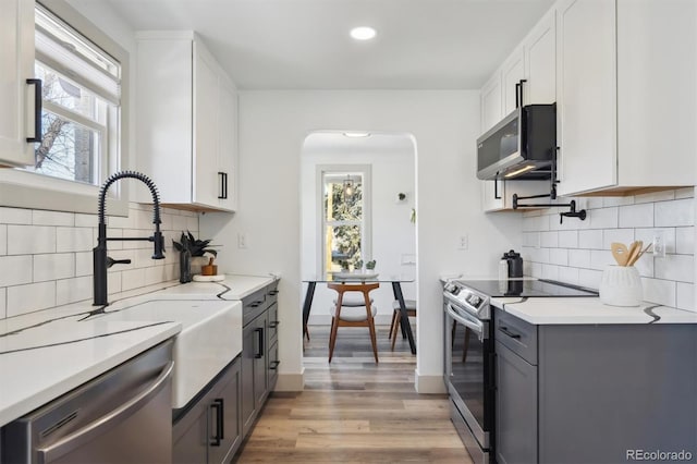 kitchen featuring arched walkways, stainless steel appliances, white cabinetry, light countertops, and light wood-type flooring