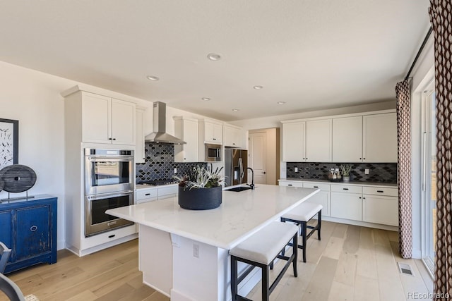 kitchen featuring a kitchen breakfast bar, wall chimney range hood, light hardwood / wood-style flooring, an island with sink, and stainless steel appliances