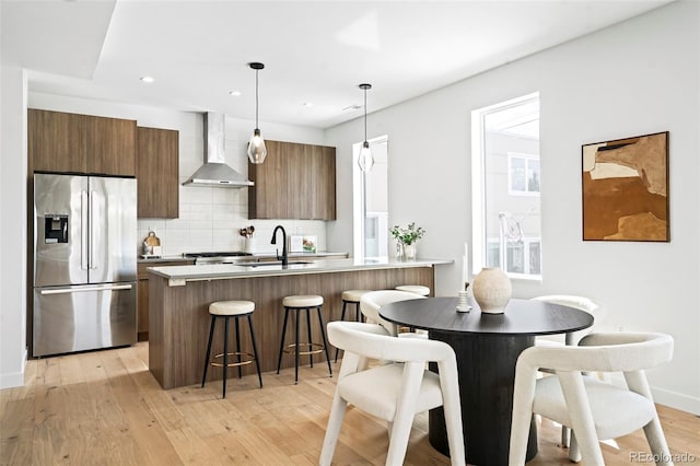 kitchen with wall chimney exhaust hood, sink, decorative light fixtures, stainless steel fridge with ice dispenser, and a center island with sink