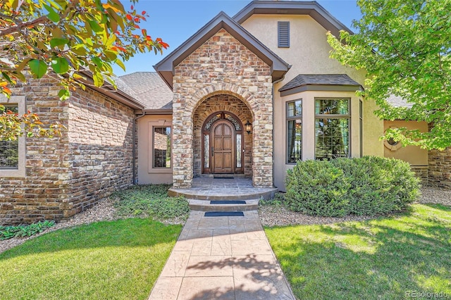 4725 Palmer Ridge Dr, Parker, CO 80134 - view of exterior entry with stucco siding, stone siding, a lawn, and a shingled roof