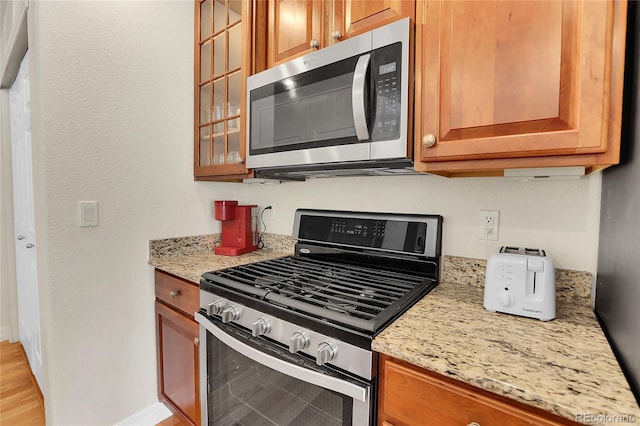 kitchen with baseboards, glass insert cabinets, brown cabinets, light stone counters, and stainless steel appliances