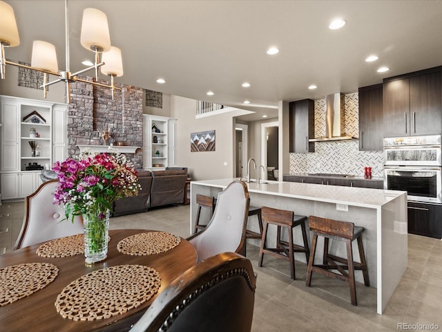 kitchen with backsplash, dark brown cabinets, wall chimney range hood, a center island with sink, and a chandelier