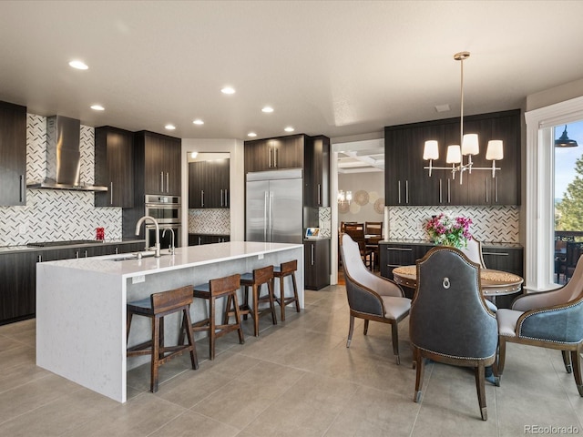 kitchen with wall chimney exhaust hood, dark brown cabinetry, a kitchen island with sink, sink, and decorative light fixtures