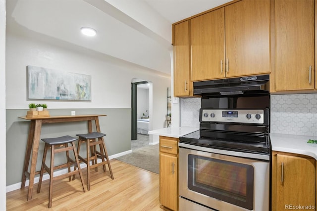 kitchen featuring stainless steel electric stove, under cabinet range hood, arched walkways, light countertops, and decorative backsplash