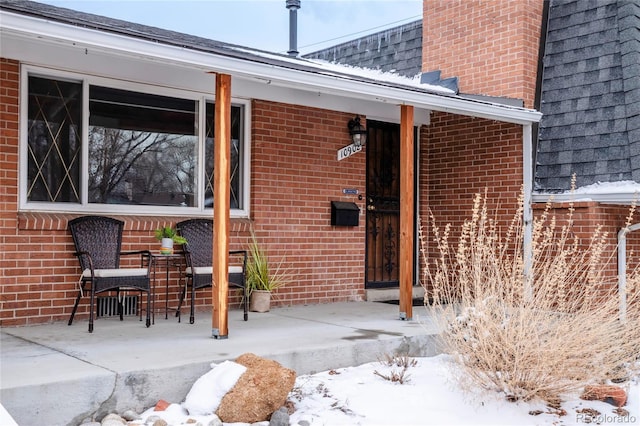 snow covered property entrance featuring brick siding and a shingled roof