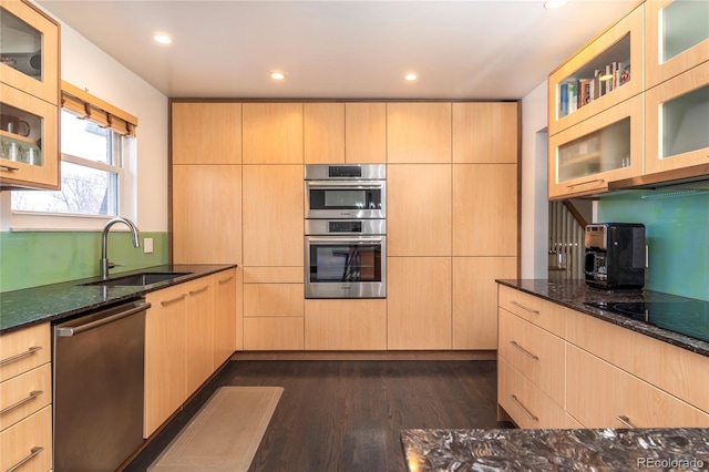 kitchen with glass insert cabinets, dark stone countertops, stainless steel appliances, light brown cabinetry, and a sink