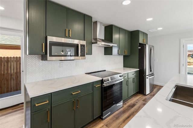 kitchen with light stone counters, wall chimney exhaust hood, stainless steel appliances, and green cabinetry