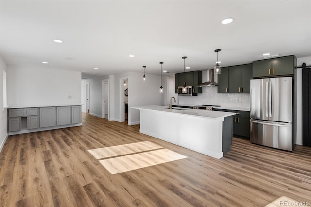 kitchen featuring pendant lighting, wall chimney exhaust hood, light wood-type flooring, a center island with sink, and stainless steel appliances