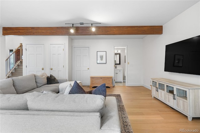living area with track lighting, beamed ceiling, light wood-style floors, baseboards, and stairway