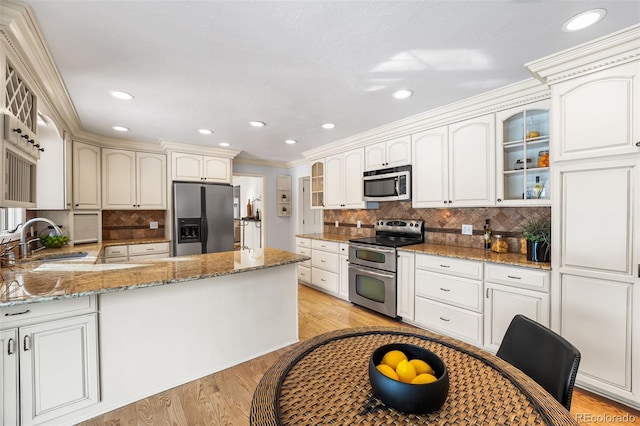 kitchen with light stone counters, a sink, appliances with stainless steel finishes, light wood finished floors, and glass insert cabinets