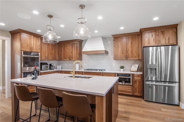 kitchen featuring sink, custom exhaust hood, appliances with stainless steel finishes, an island with sink, and pendant lighting