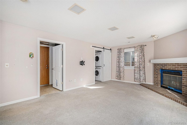 unfurnished living room featuring a brick fireplace, stacked washer and clothes dryer, light colored carpet, and a barn door