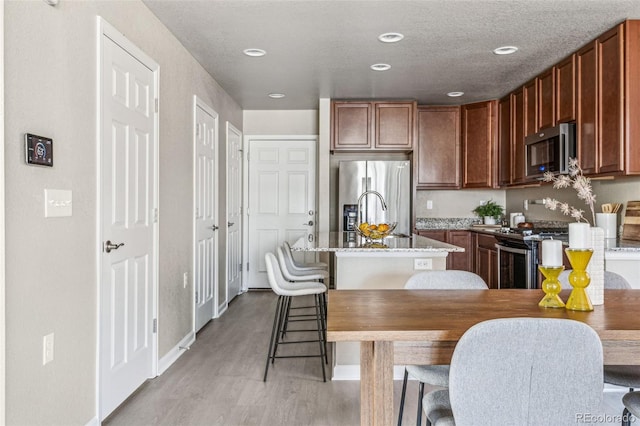 kitchen with appliances with stainless steel finishes, a sink, a textured ceiling, an island with sink, and light wood-type flooring