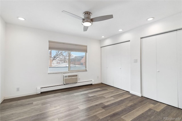 unfurnished bedroom featuring an AC wall unit, baseboard heating, two closets, hardwood / wood-style flooring, and ceiling fan