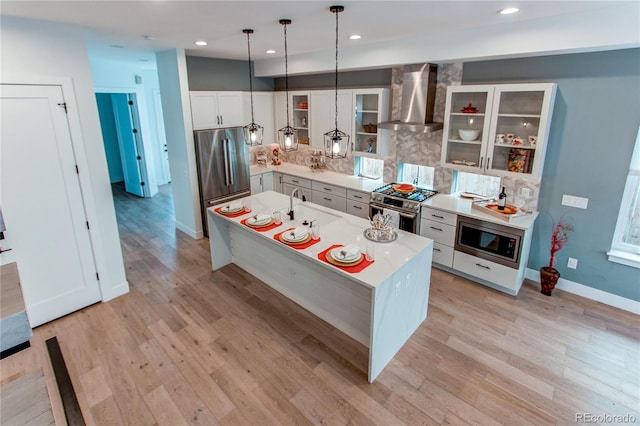 kitchen featuring an island with sink, decorative light fixtures, light hardwood / wood-style floors, appliances with stainless steel finishes, and wall chimney range hood