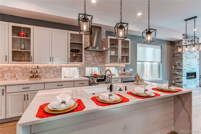 kitchen featuring a center island with sink, sink, white cabinetry, decorative light fixtures, and wall chimney range hood